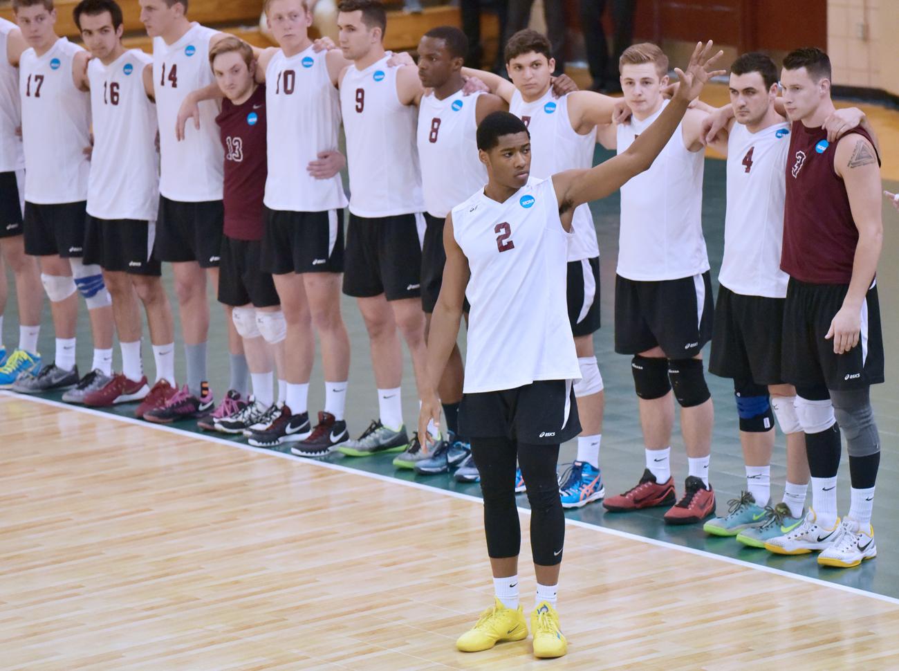 Greg Woods in his volleyball uniform standing on the court waving to the crowd, there are several of his teammates standing in a line behind him